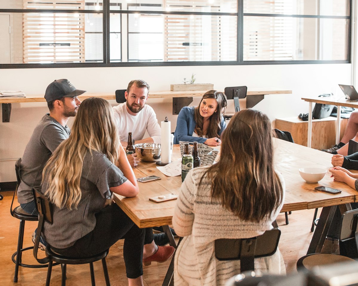 Six people talk around a long wooden table in a bright room.