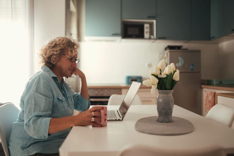 A woman sits at a bright kitchen table with a laptop and a cup of tea.
